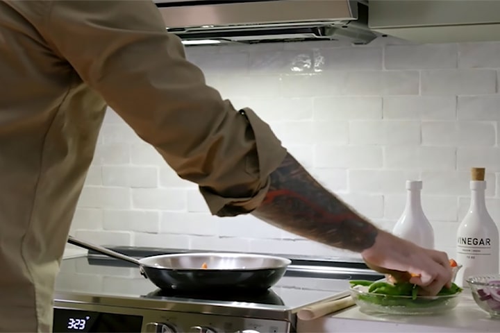 A person with a tattooed arm cooking vegetables in a stainless steel frying pan on a modern glass stovetop.